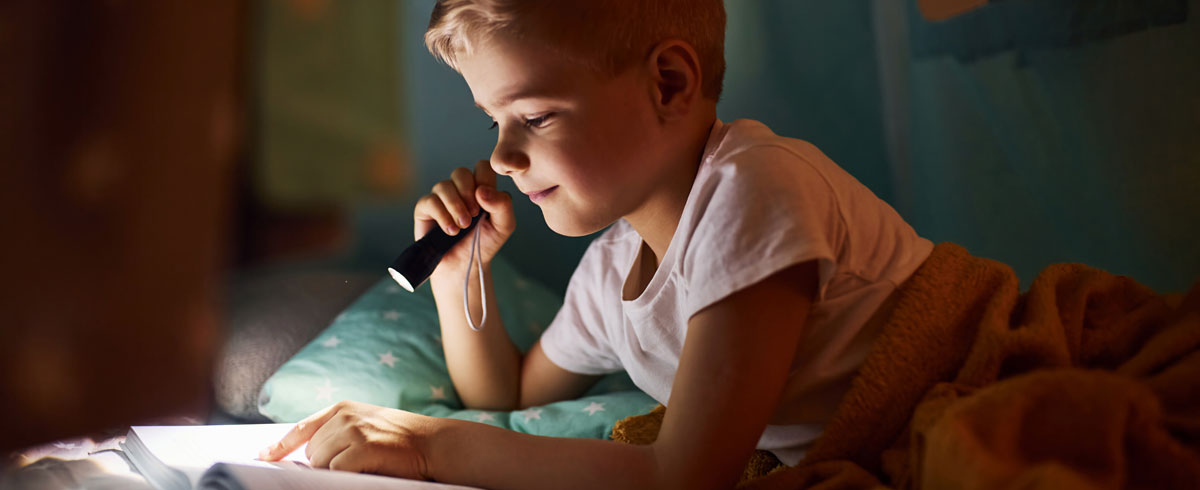 Un bambino che legge un libro con una torcia elettrica in mano mentre è nel letto con una coperta